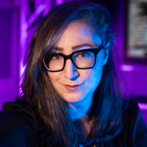 headshot of a white woman with medium length straight brown hair and black rectangular glasses, with a background showing a speaker and a monitor with code on it awash in purple light